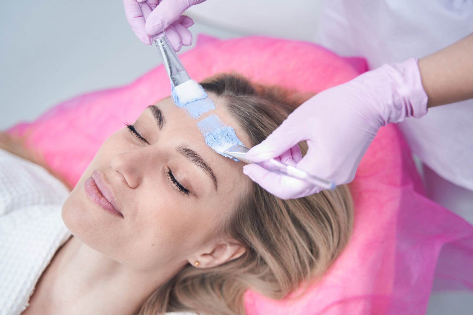 Cosmetologist applying mask on female client skin Cosmetician hands spreading blue clay over woman forehead with two cosmetic brushes