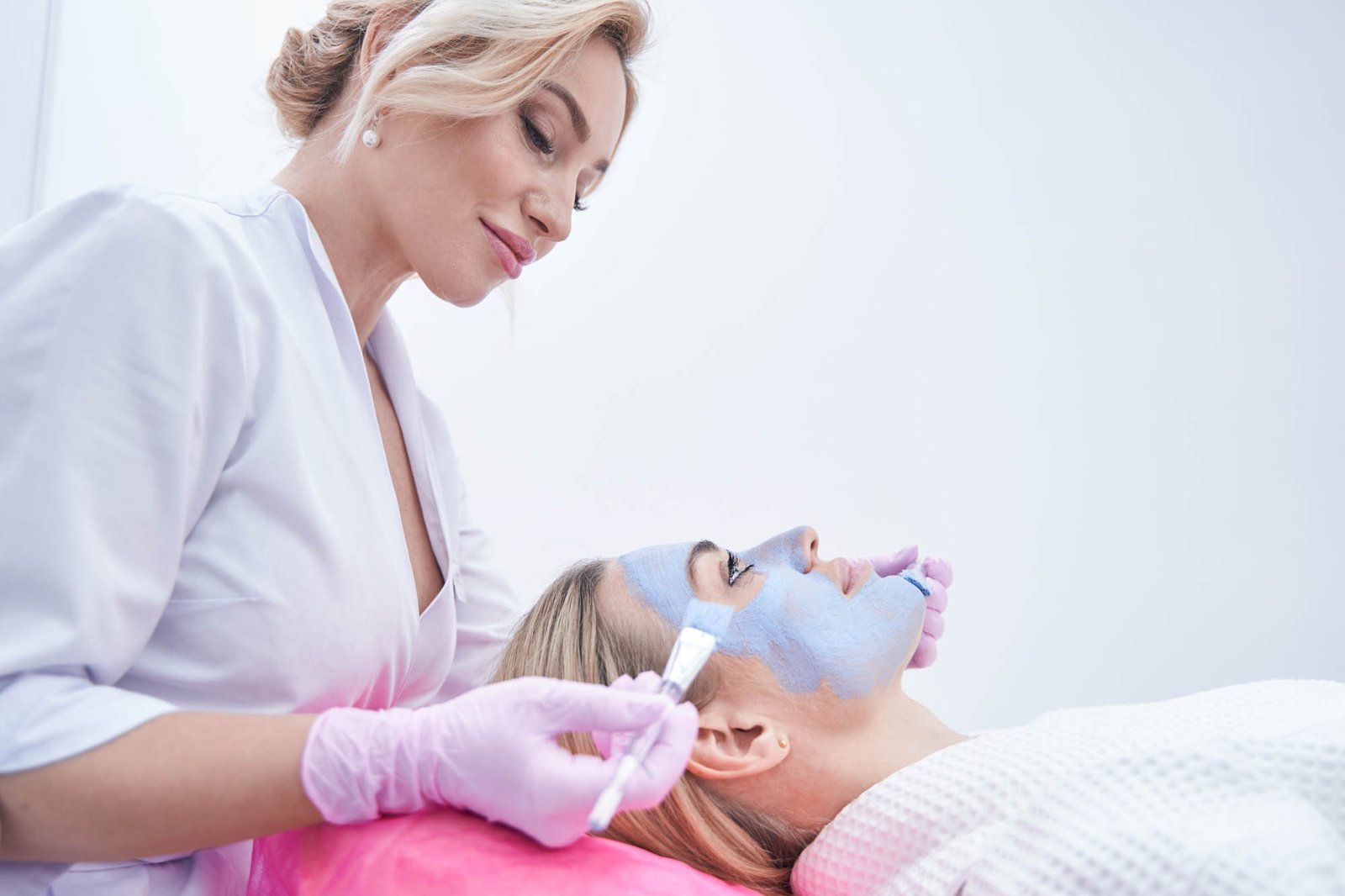 Focused beautician applying mask to face of calm young woman Cosmetician spreading blue clay paste over facial skin of client using two brushes